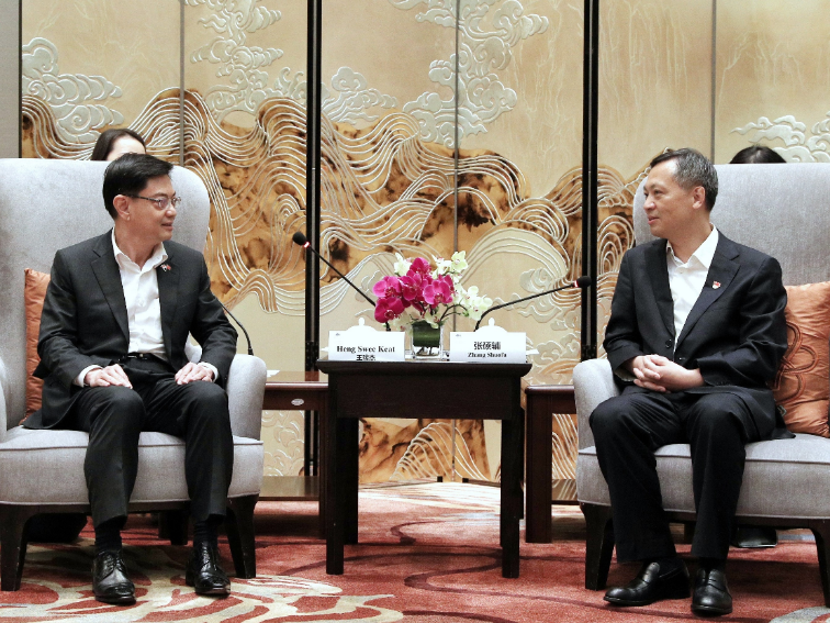 Two men in suits seated in chairs, facing each other with table between them, with name cards present.